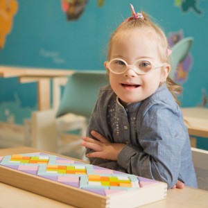 smiling girl with glasses sitting at a classroom desk. A box of pattern blocks are in front of her.