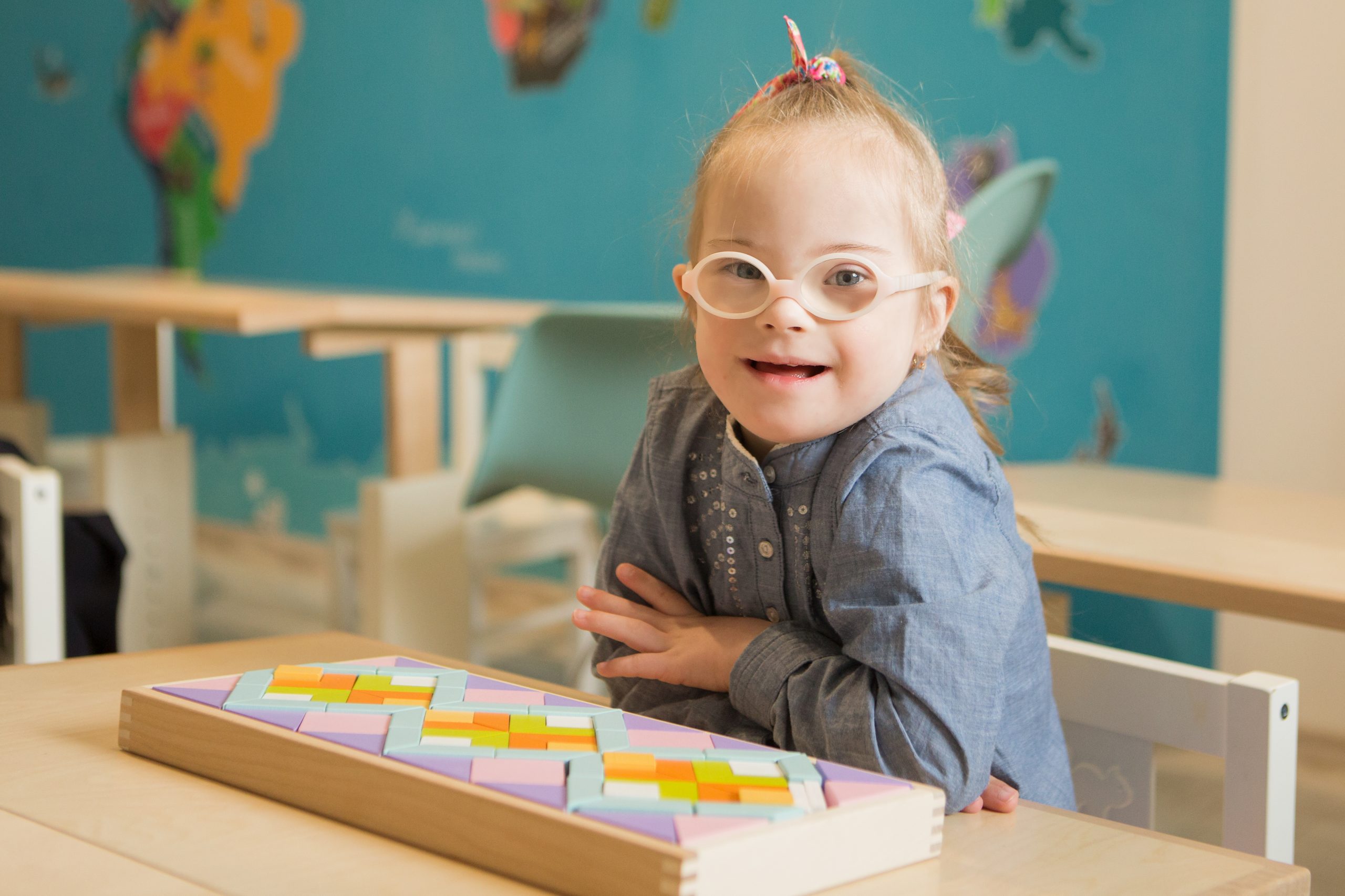 smiling girl with glasses sitting at a classroom desk. A box of pattern blocks are in front of her.