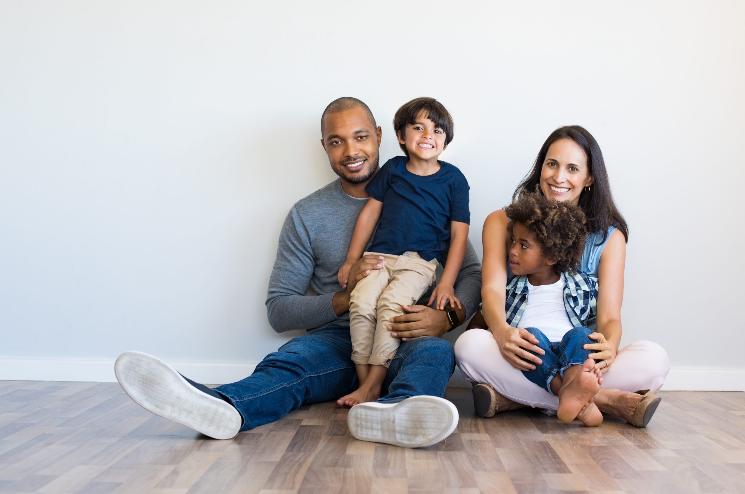 A smiling family sits on the floor while holding their two children on their laps.