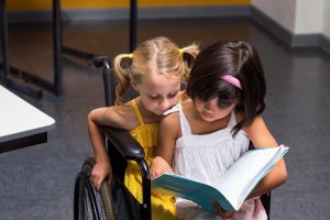 Two young girls read a book together. One girl is in a wheelchair and the other shares the seat while they look at the book.